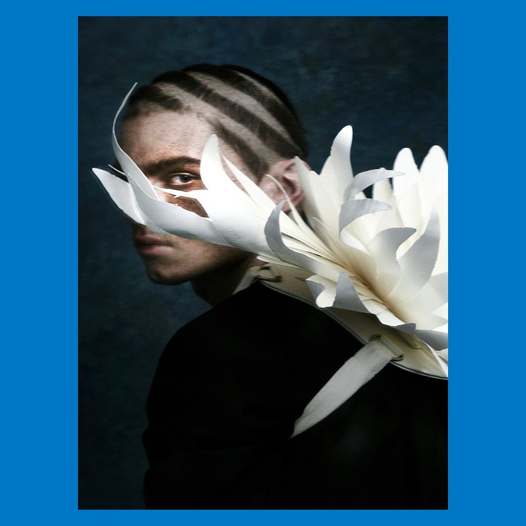 Artistic portrait of a male model wearing a unique white paper floral collar and bold striped hairstyle against a dark backdrop.