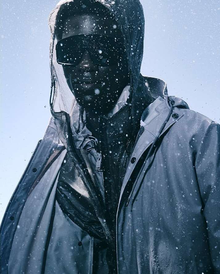 Man wearing a hooded raincoat and dark sunglasses under falling water droplets against a clear sky background