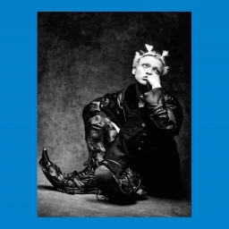 Monochrome portrait of a seated person with spiky hair wearing a leather jacket and oversized pointed boots against a textured backdrop