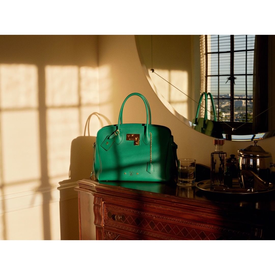 Elegant green leather handbag on a dark wood dresser with ornate carvings, illuminated by warm sunlight in a sophisticated interior.