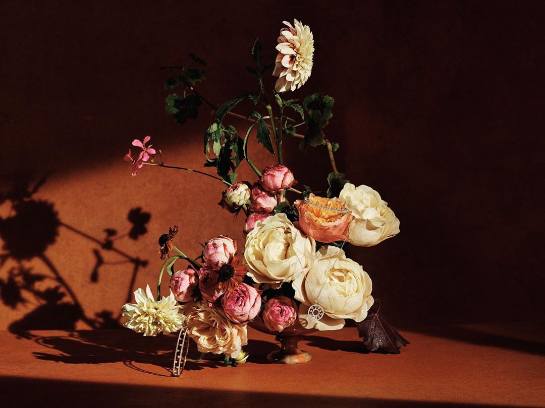 Dramatic floral arrangement featuring roses and dahlias in warm, muted tones, set against a textured, earthy background.
