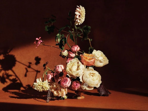Dramatic floral arrangement featuring roses and dahlias in warm, muted tones, set against a textured, earthy background.