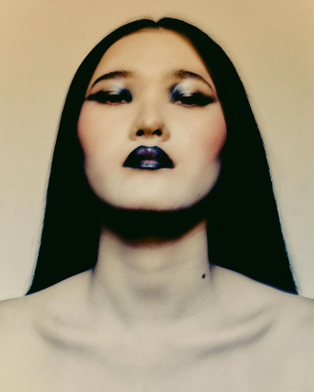 Close-up portrait of a woman with dark hair, blue eye makeup, and dark lipstick against a plain background