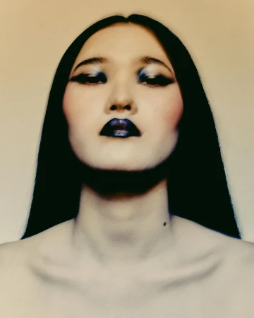 Close-up portrait of a woman with dark hair, blue eye makeup, and dark lipstick against a plain background