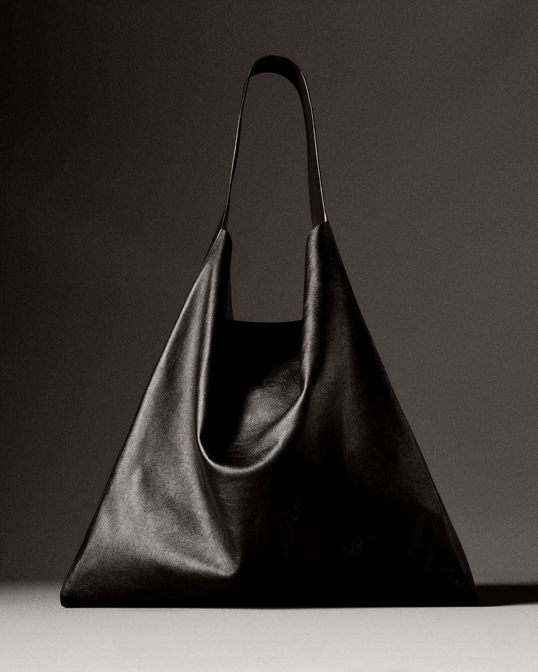 Studio shot of a black leather tote bag with a triangular shape and long handles against a dark gray background with soft lighting.