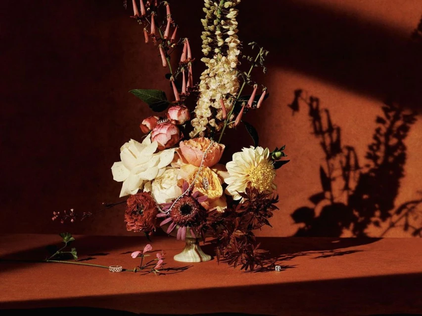 Floral arrangement featuring a mix of roses, dahlias, and wildflowers in a vase on a textured reddish-orange surface with dramatic lighting.