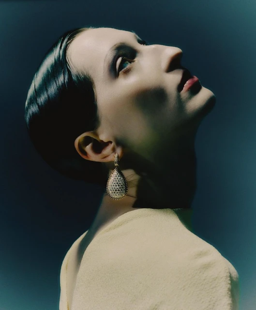 Close-up of a stylish adult female with slicked-back hair and teardrop earrings, gazing upwards against a blurred blue background.