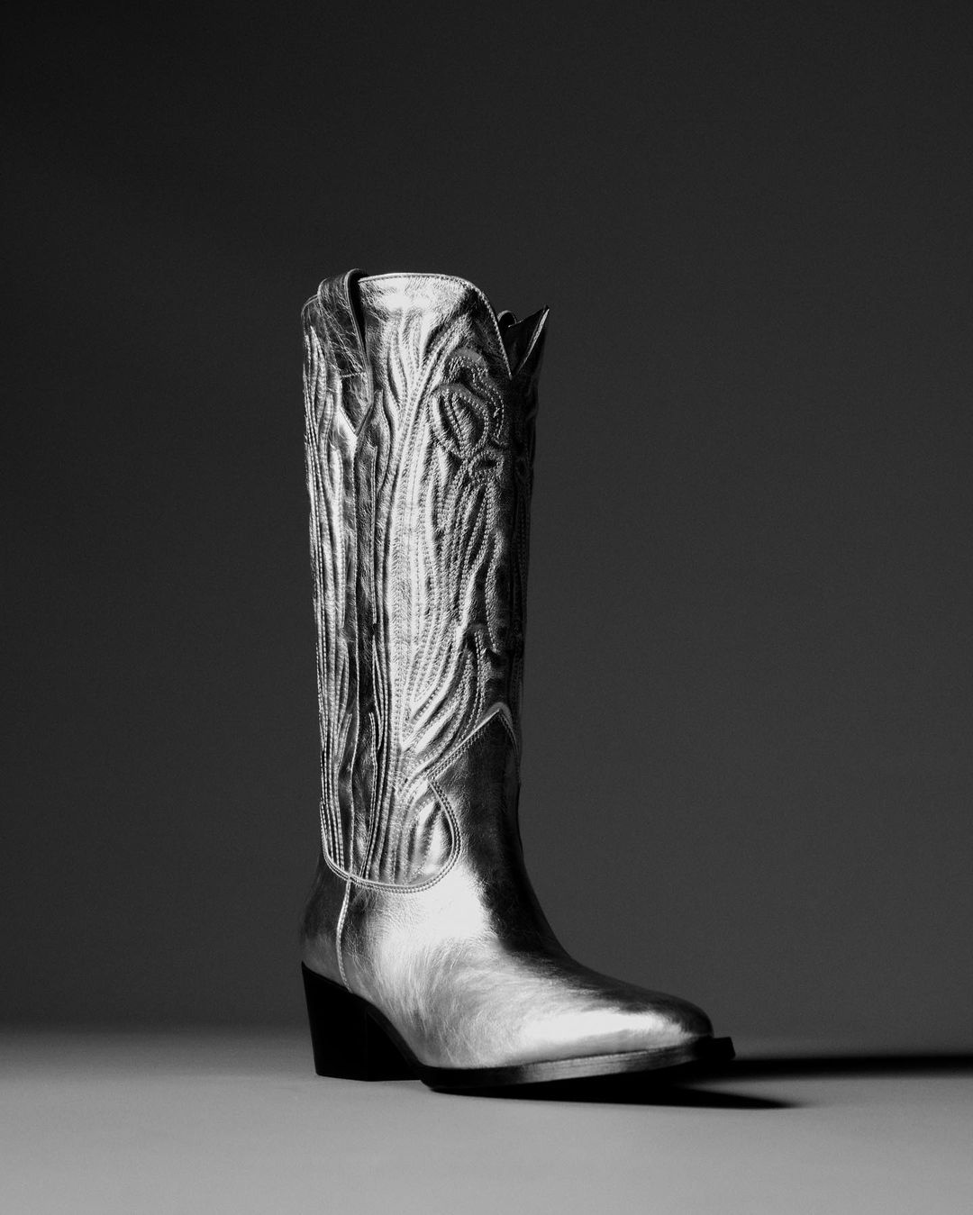 Shiny metallic cowboy boot stands tall against a neutral gray backdrop, showcasing texture and design in a studio setting.