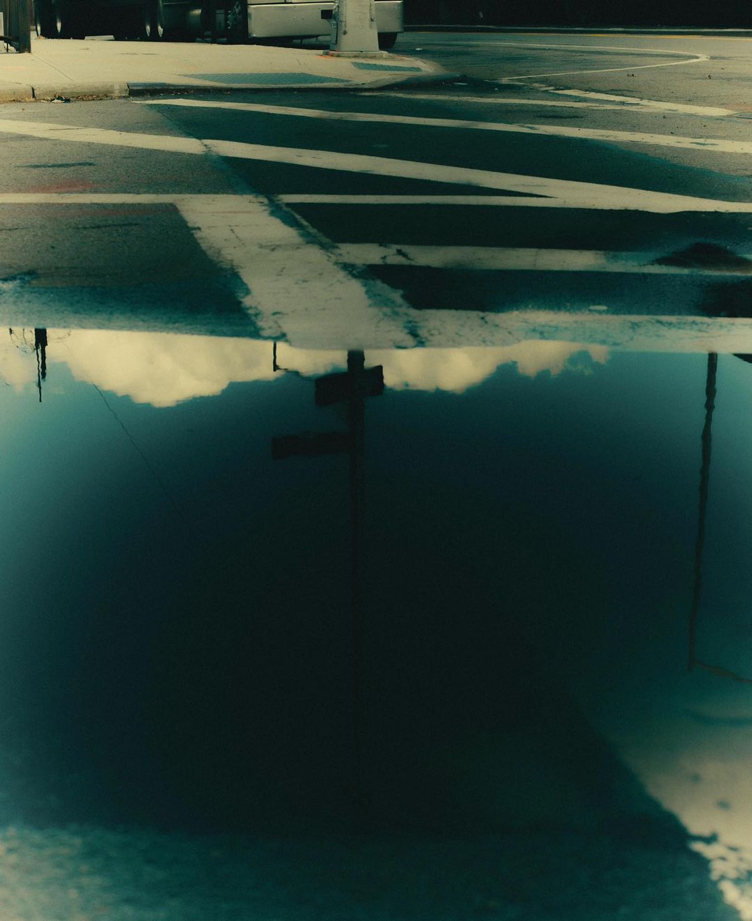 Street scene with crosswalk reflection in a puddle. Urban scene shows asphalt road with white stripes, and sky and street pole reflection.