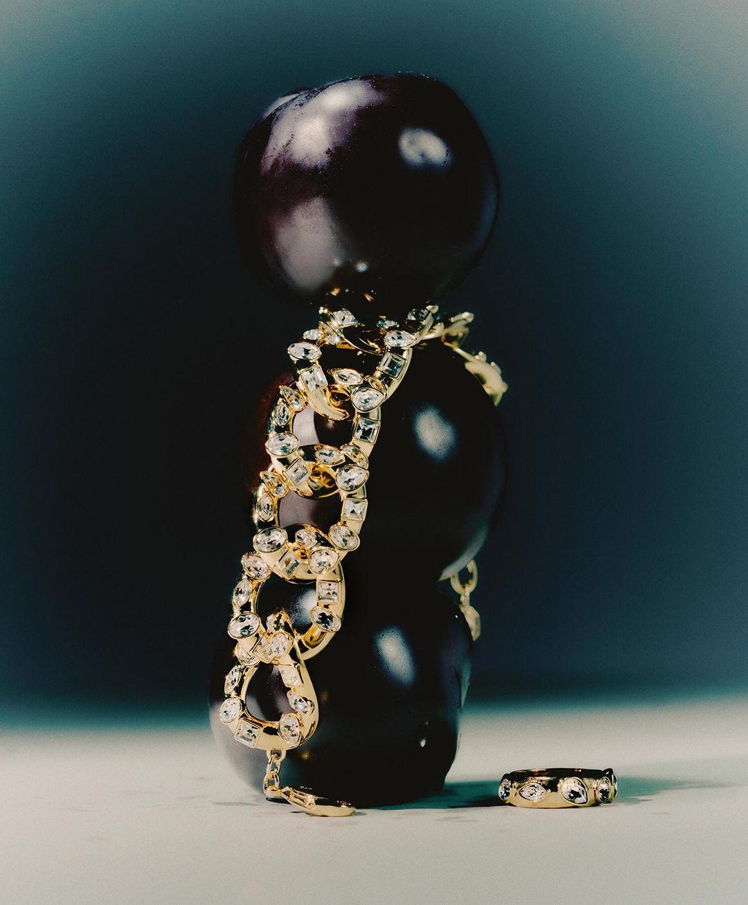Still life of three plums stacked vertically, adorned with a gold and crystal chain and a matching ring, against a dark background.