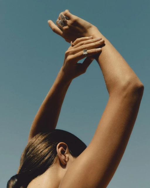Woman with arms raised wearing rings in a sunny outdoor setting, shot with a clear blue sky background. Jewelry, fashion, and summer concept.