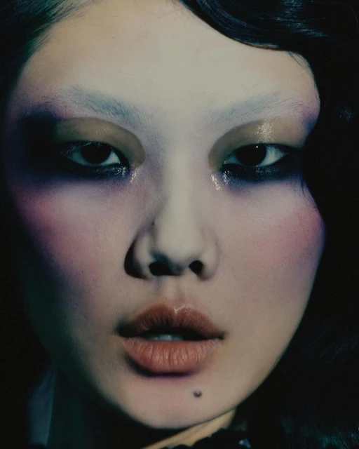Close-up portrait of a woman with pale skin and distinctive makeup featuring bold eye shadow and lip color, set against a dark backdrop.