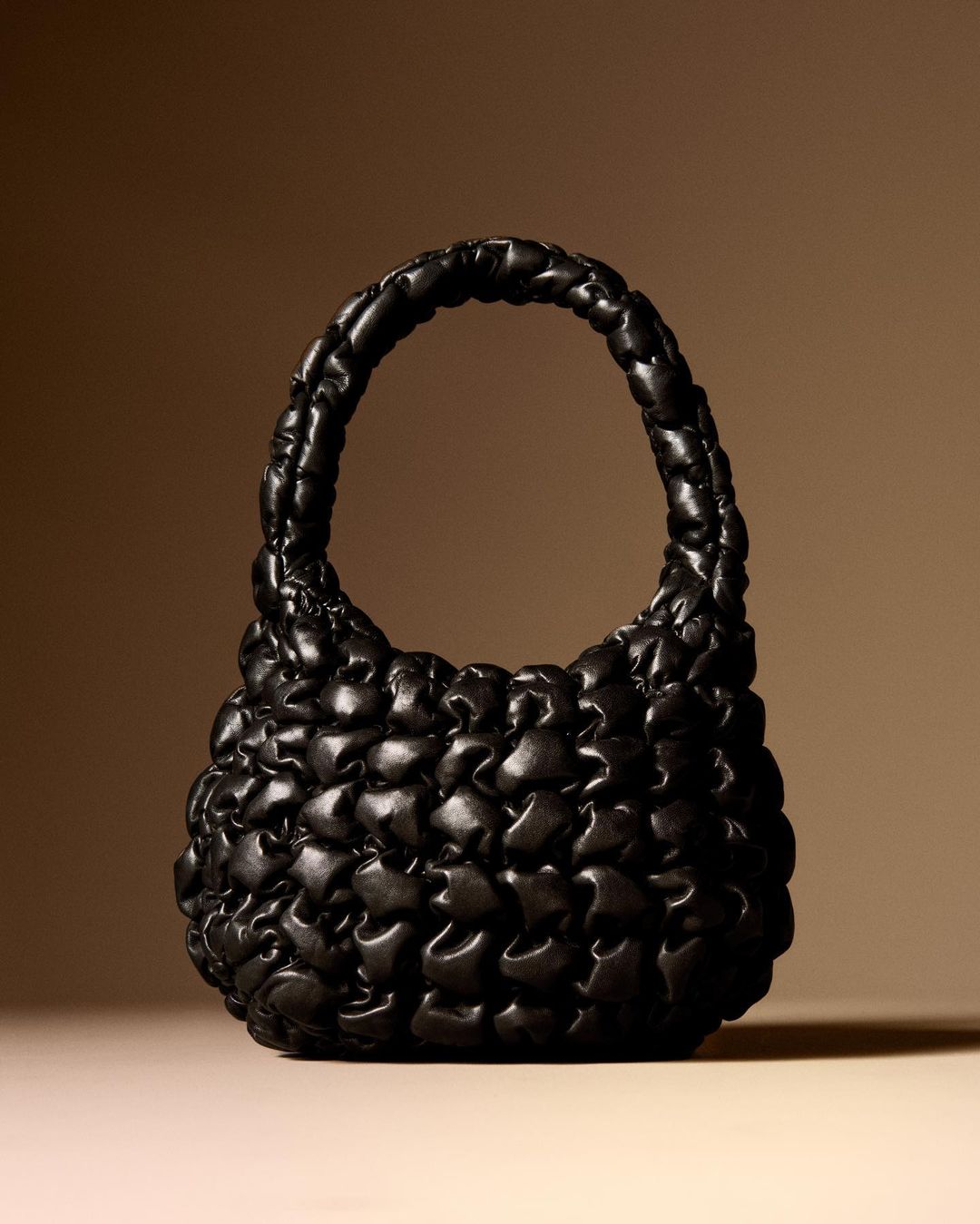Padded black leather handbag with top handle on beige background. Fashionable accessory with unique puffy design and soft texture.
