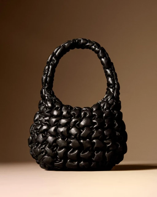 Padded black leather handbag with top handle on beige background. Fashionable accessory with unique puffy design and soft texture.