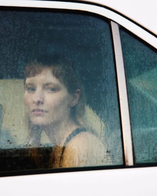 Portrait of a woman with short hair inside of a car, looking out of a rain-covered car window. The photo is shot in natural lighting.