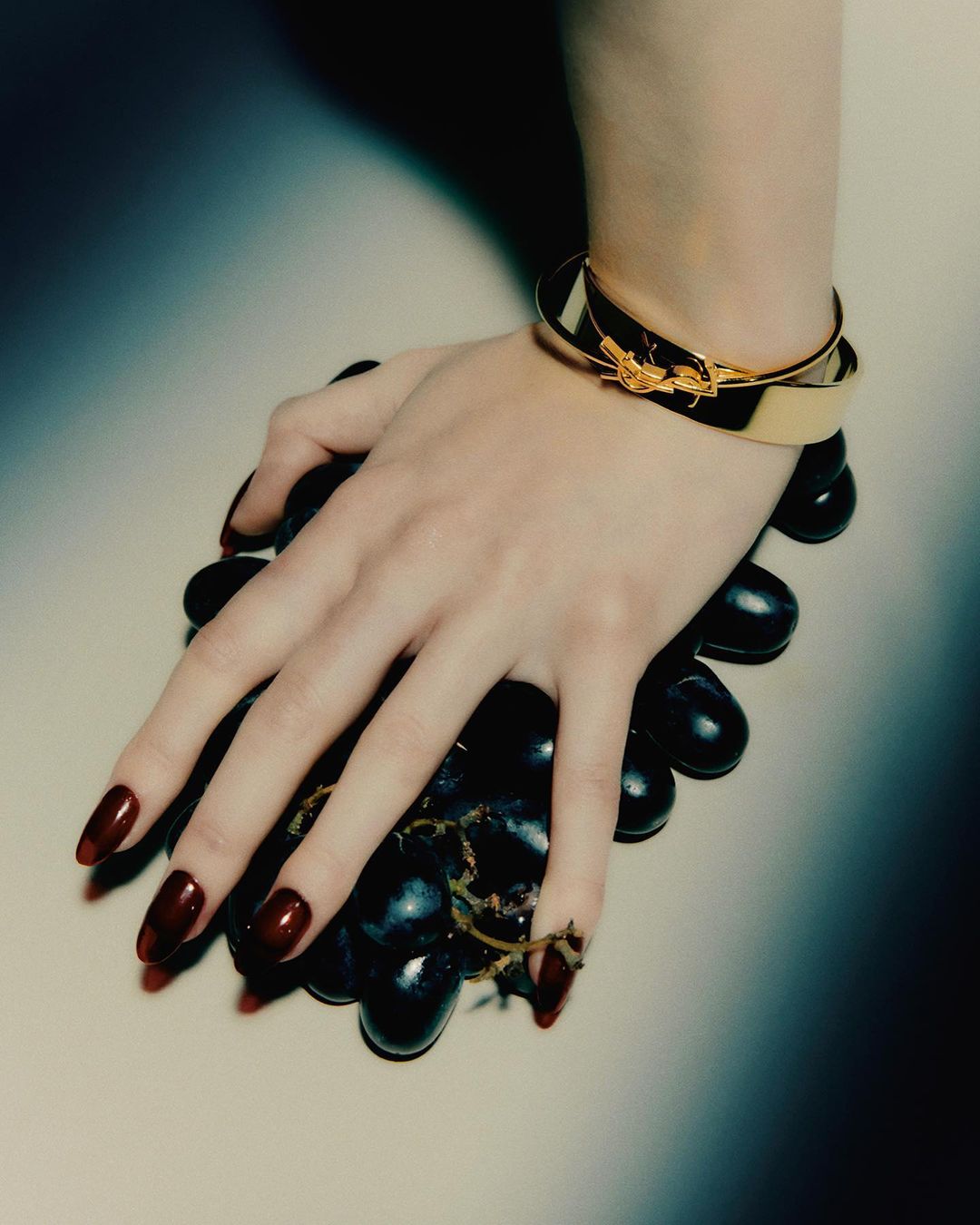 Close-up of an adult female's hand with long burgundy nails and a gold cuff bracelet holding a cluster of dark purple grapes.