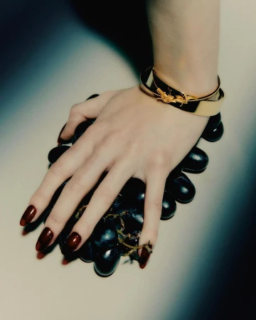 Close-up of an adult female's hand with long burgundy nails and a gold cuff bracelet holding a cluster of dark purple grapes.