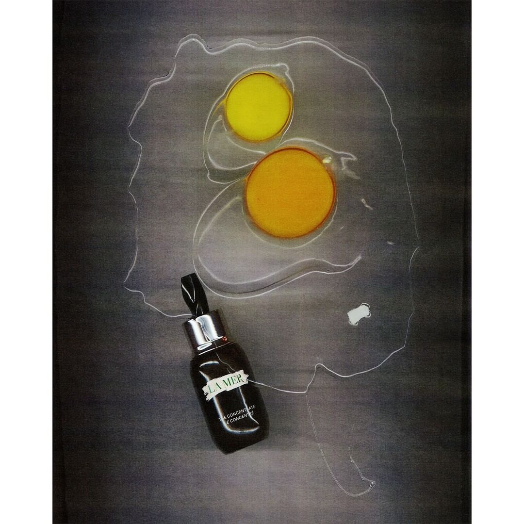 Beauty still life featuring La Mer serum bottle, yellow and orange circles, and a clear, egg-shaped outline on a dark surface.