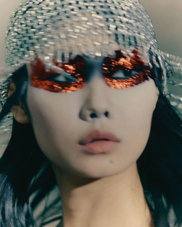 Woman with sparkling silver headpiece and glittery orange eye makeup looking off-camera with neutral expression and dark hair.