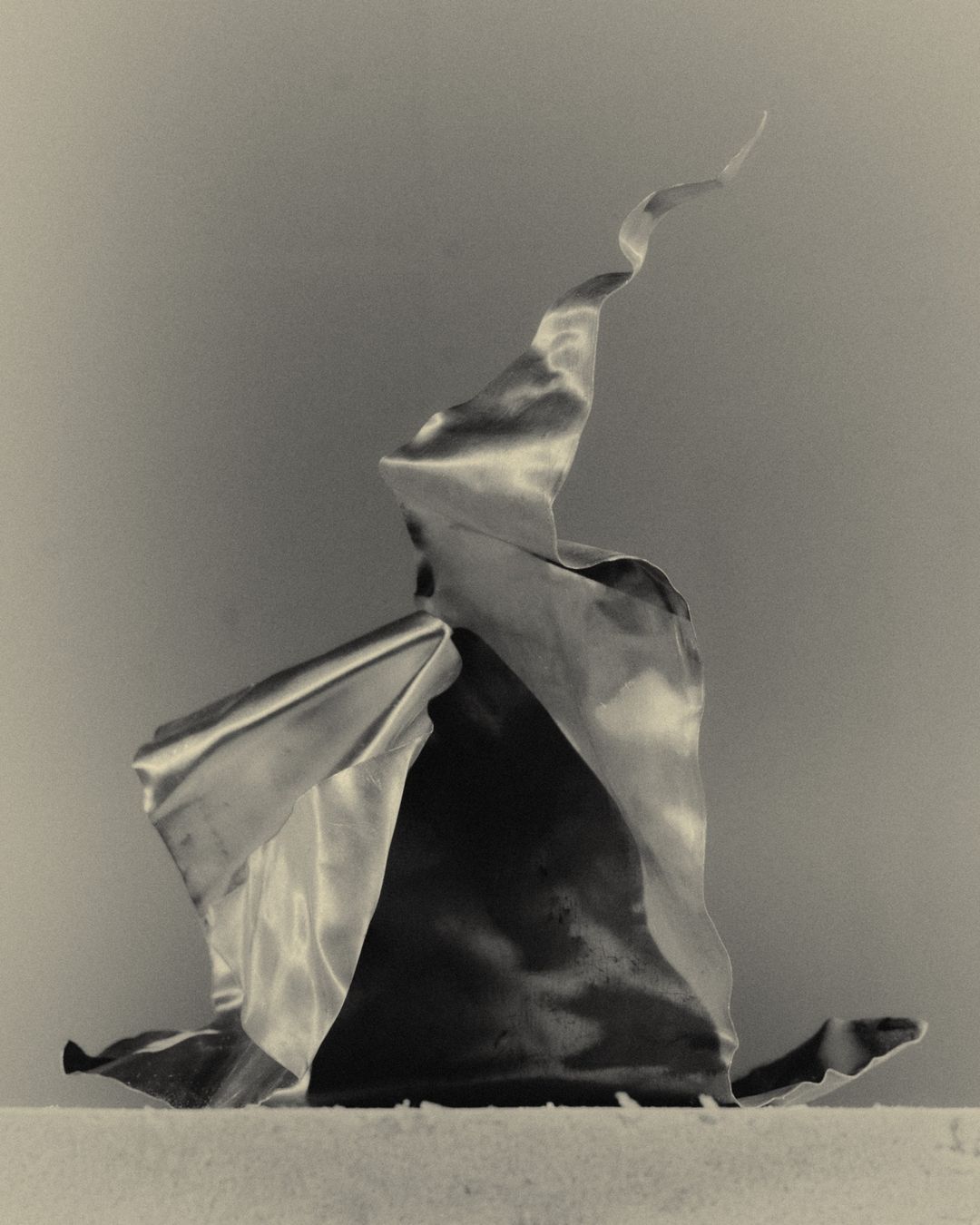 Abstract studio photograph of a wavy, reflective satin ribbon against a gradient grey background. The monochrome composition emphasizes form and texture.