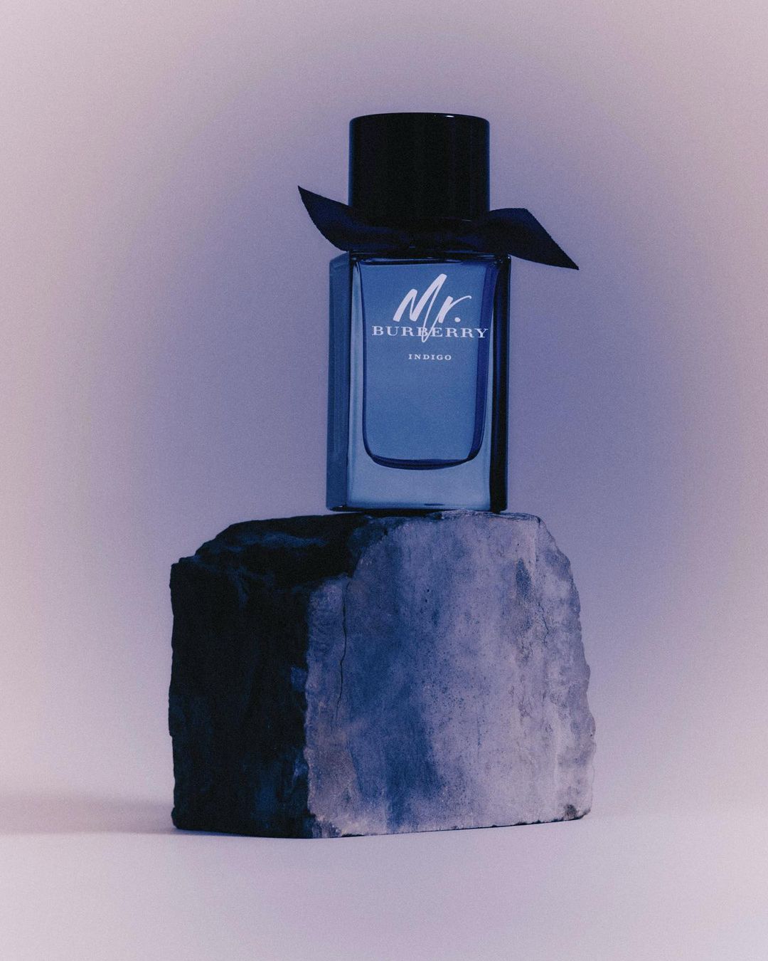 Mr. Burberry Indigo perfume bottle stands atop a textured gray stone, set against a gradient backdrop for a beauty product ad.