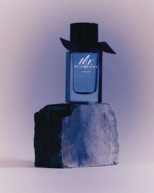 Mr. Burberry Indigo perfume bottle stands atop a textured gray stone, set against a gradient backdrop for a beauty product ad.