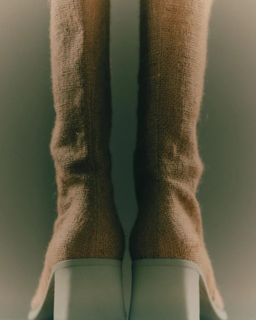Close-up studio shot of tan textile boots with white block heels. The boots are set against a soft, diffused grey background.