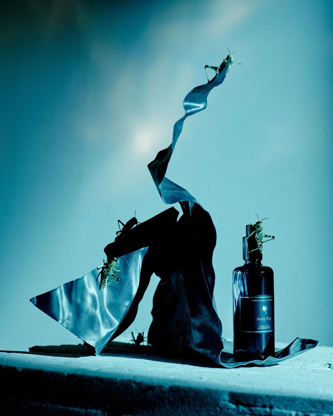 Dark perfume bottle with insects, flowing fabric and teal lighting. Product shot for fragrance brand with surreal presentation.
