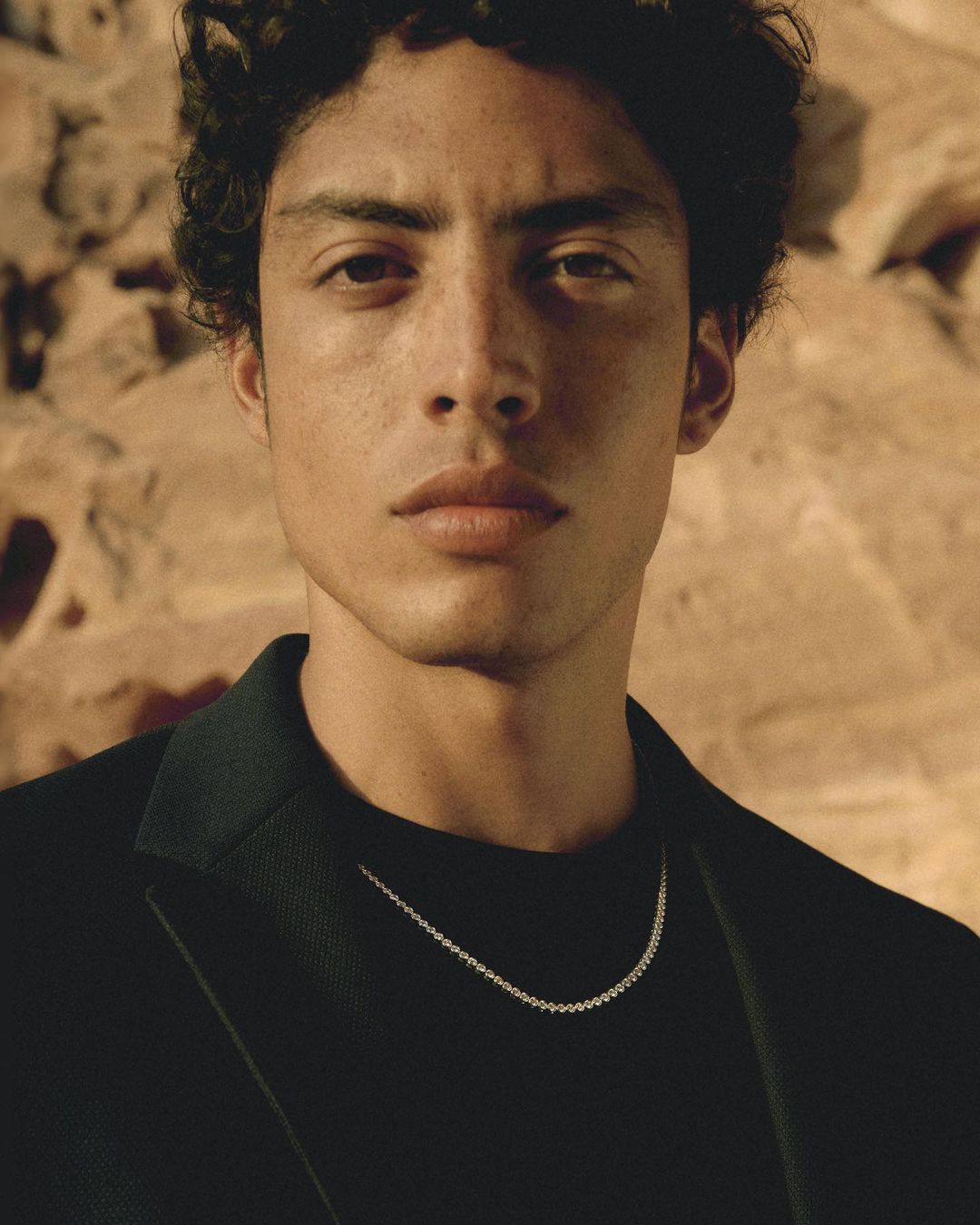 Close-up portrait of a young man wearing a black blazer and necklace against a rocky earth background.