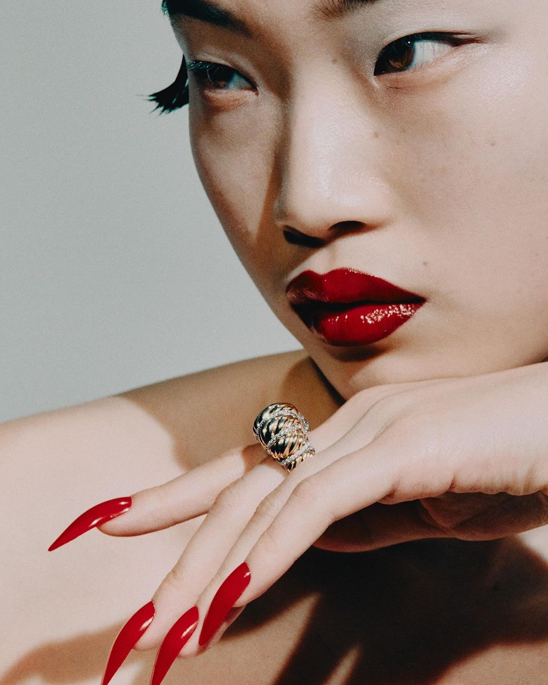 Close-up of an Asian woman with red lipstick and long red nails, wearing an ornate gold ring, against a light gray background.
