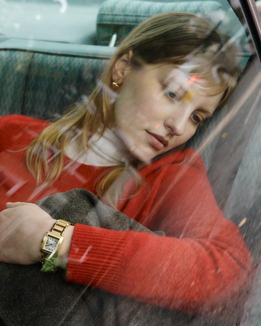 Portrait of a blonde woman inside a vehicle, wearing a red sweater and gold watch, glancing downward with a pensive expression.