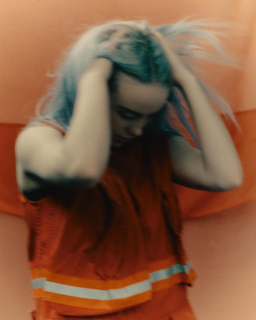 A blurry medium shot of a young adult female with blue hair in an orange reflective vest, touching her hair, set against a blurry orange background.