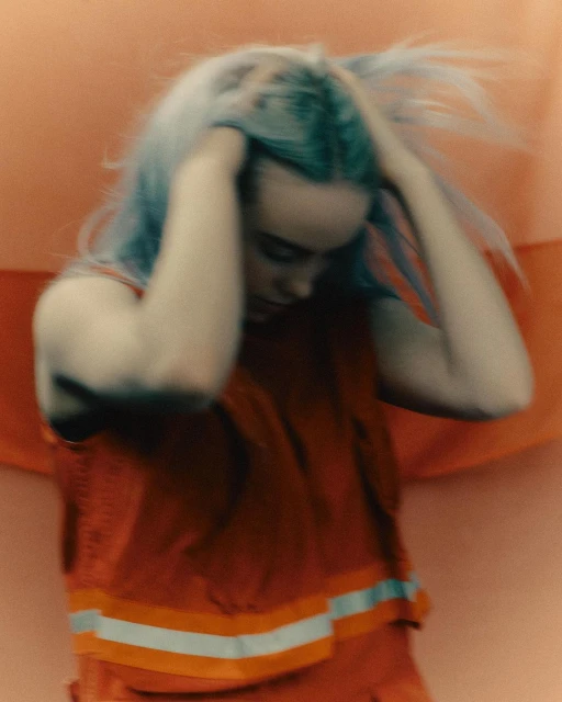 A blurry medium shot of a young adult female with blue hair in an orange reflective vest, touching her hair, set against a blurry orange background.