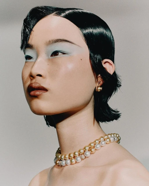 Close-up of a young Asian woman with a stylish bob haircut, wearing elegant gold and white pearl jewelry and light blue eyeshadow.