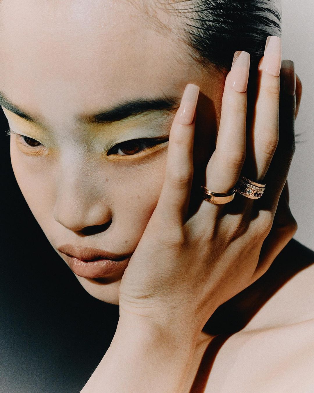 Close-up portrait of a young woman with yellow eyeshadow and rings, her face softly lit against a partially dark background.