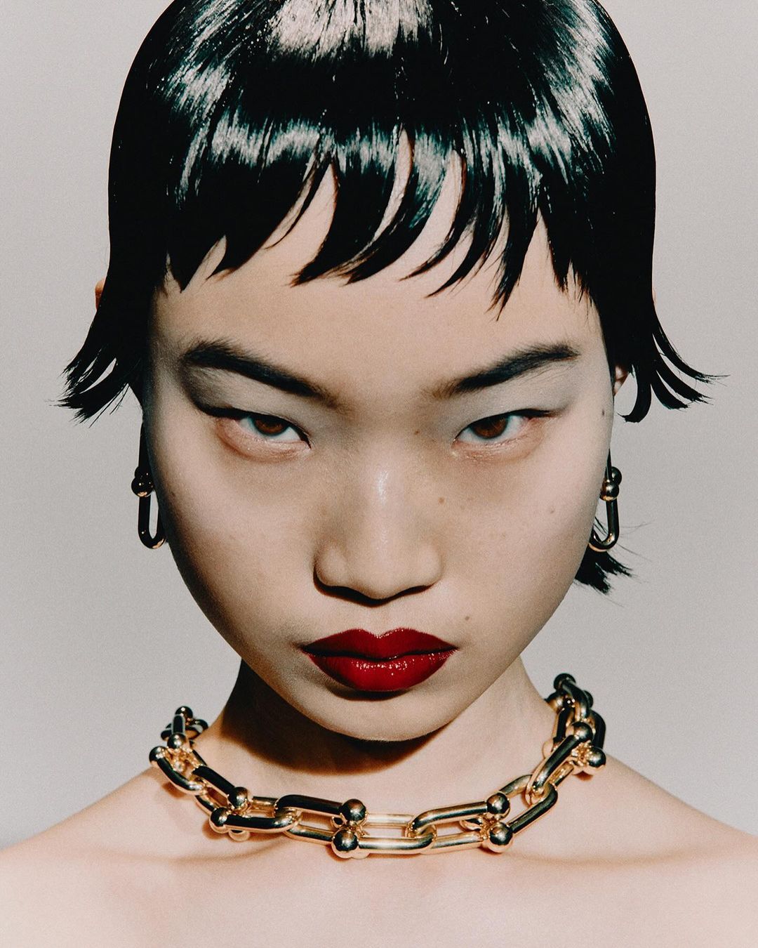 Close-up portrait of a model with dark wet-look fringe hair, glossy red lips, gold chain necklace, and hoop earrings against a light background.