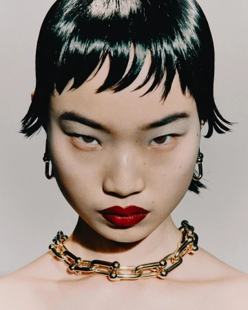 Close-up portrait of a model with dark wet-look fringe hair, glossy red lips, gold chain necklace, and hoop earrings against a light background.