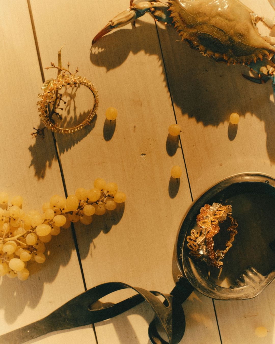 Overhead shot featuring a blue crab, grapes, decorative hoop, and metal utensils on a white wooden surface with strong directional lighting.