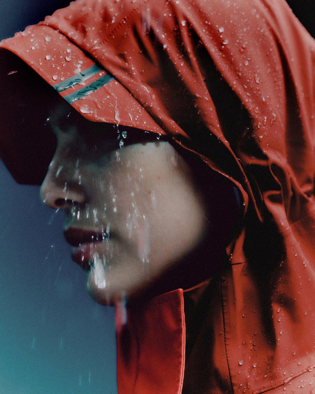 Close-up profile of a woman wearing a red hooded rain jacket with water droplets running down her face and clothing.