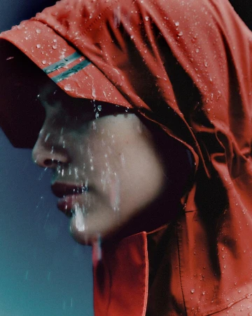 Close-up profile of a woman wearing a red hooded rain jacket with water droplets running down her face and clothing.