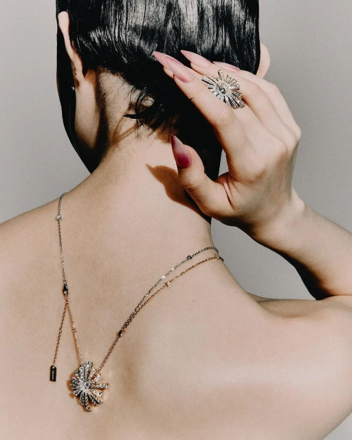 Close-up from behind of a woman with short dark hair, wearing a sparkling ring, a delicate necklace and pink manicured nails.