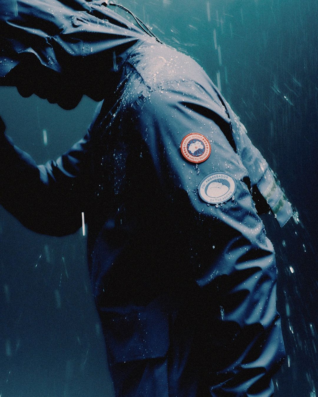Person in a dark blue hooded rain jacket with logos in the rain, creating a somber, wet atmosphere and highlighting waterproof apparel.