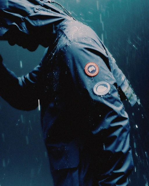 Person in a dark blue hooded rain jacket with logos in the rain, creating a somber, wet atmosphere and highlighting waterproof apparel.