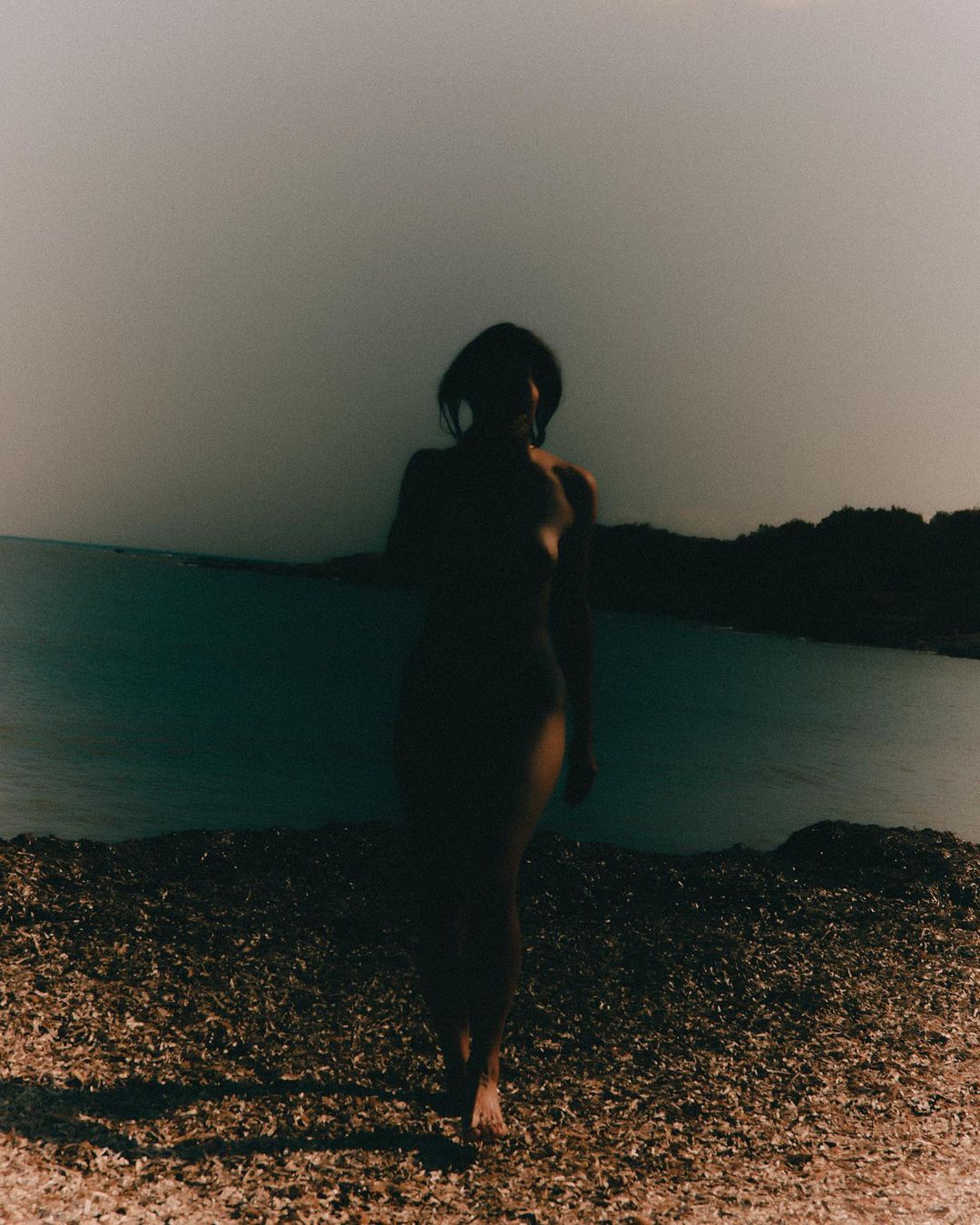 A woman walks along a sandy beach next to a body of water. The photo is desaturated and gives a peaceful and natural feeling.