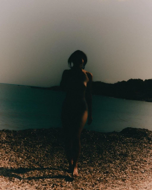A woman walks along a sandy beach next to a body of water. The photo is desaturated and gives a peaceful and natural feeling.
