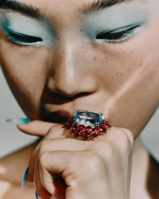Close-up of a woman's face with striking light blue eye shadow, showcasing a large cocktail ring with blue and red gemstones.
