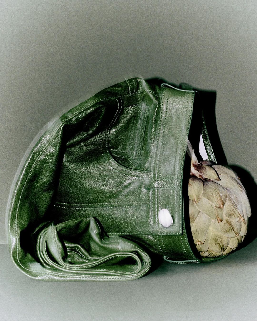 Green leather jacket still life with an artichoke, arranged on a grey backdrop, showcasing textures and muted color palette.
