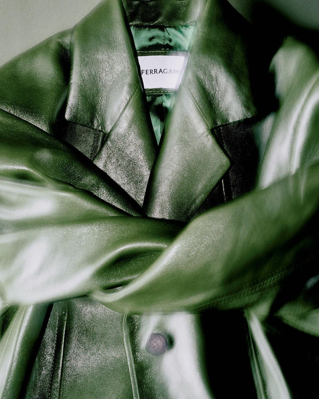 Close-up of a draped, olive green leather jacket with a 'Ferragamo' label, showcasing luxury fashion and craftsmanship in soft lighting.