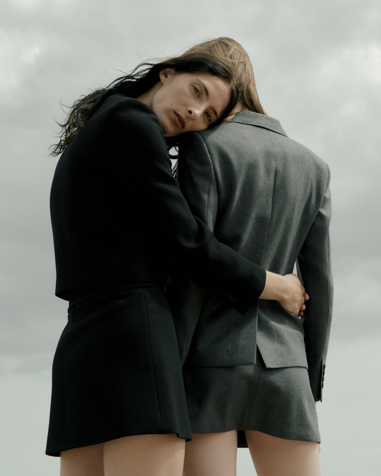 Two women embrace outdoors, wearing matching black and gray skirt suits against a muted sky, showcasing minimalist fashion.