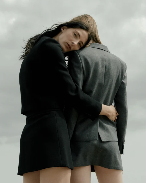 Two women embrace outdoors, wearing matching black and gray skirt suits against a muted sky, showcasing minimalist fashion.